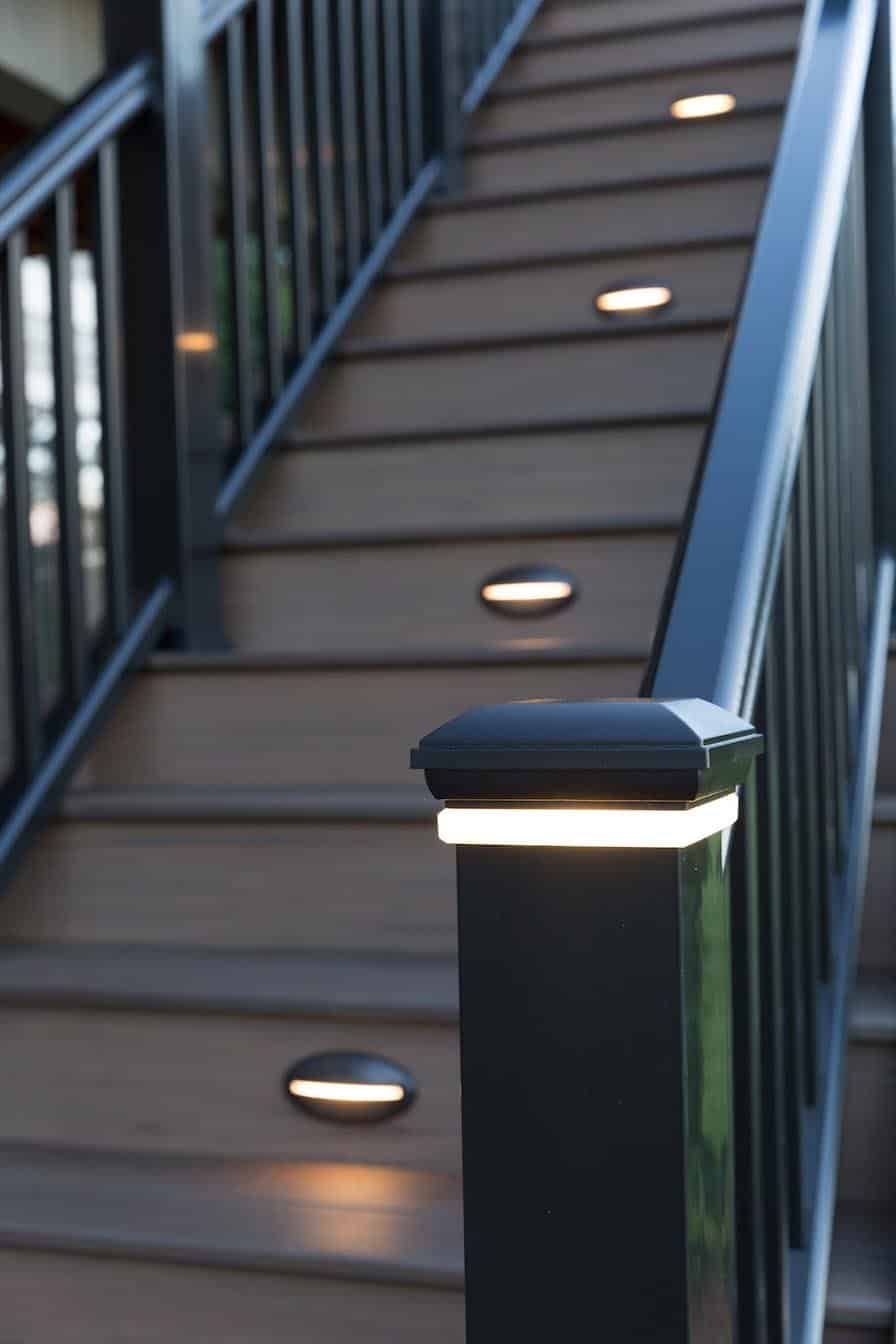 Post cap light mounted on a railing next to illuminated riser lights on a staircase.