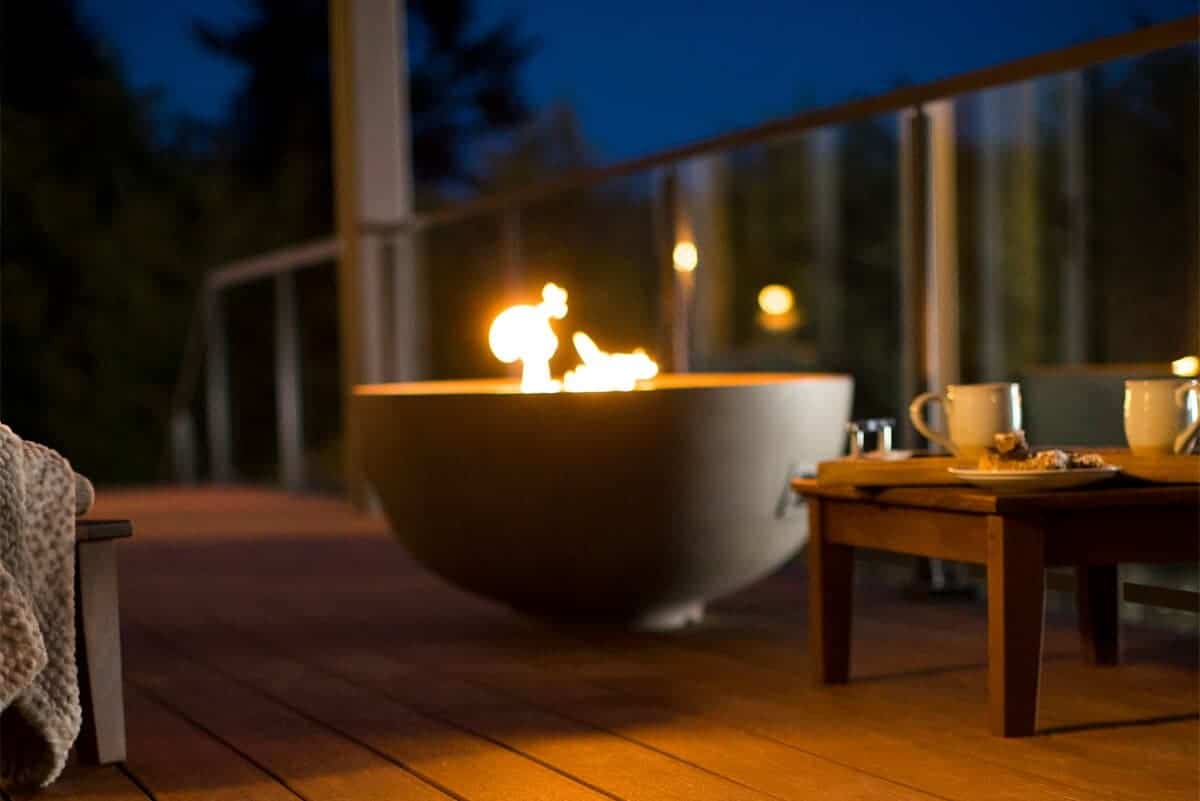 A modern outdoor fire pit with flames visible, set on a wooden deck at night.