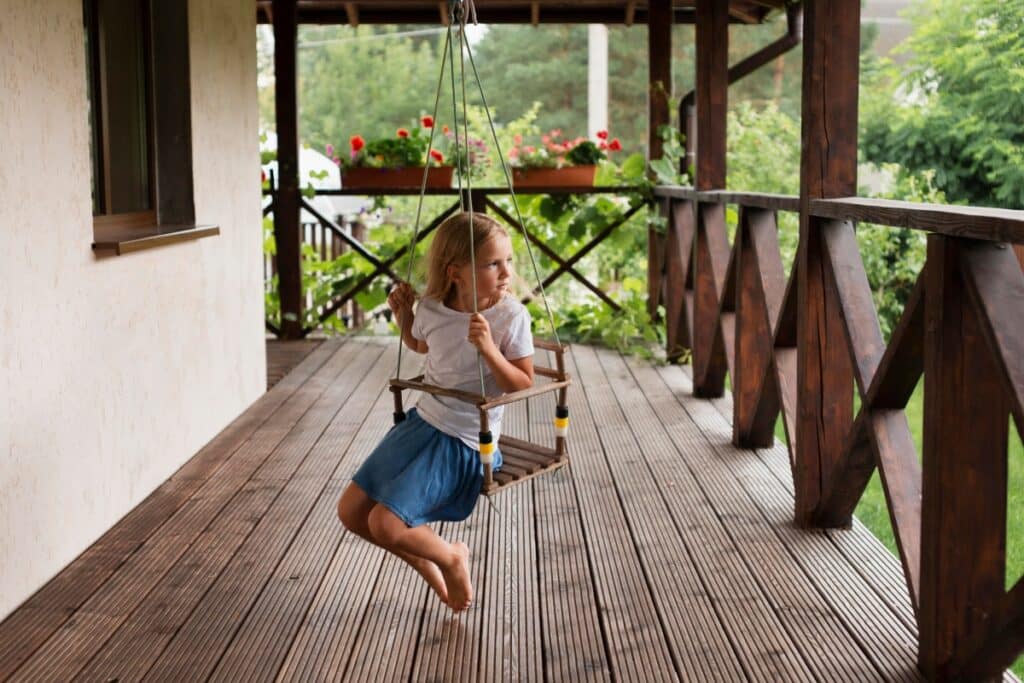 A little girl swinging in a wooden swing on a composite deck around the house.